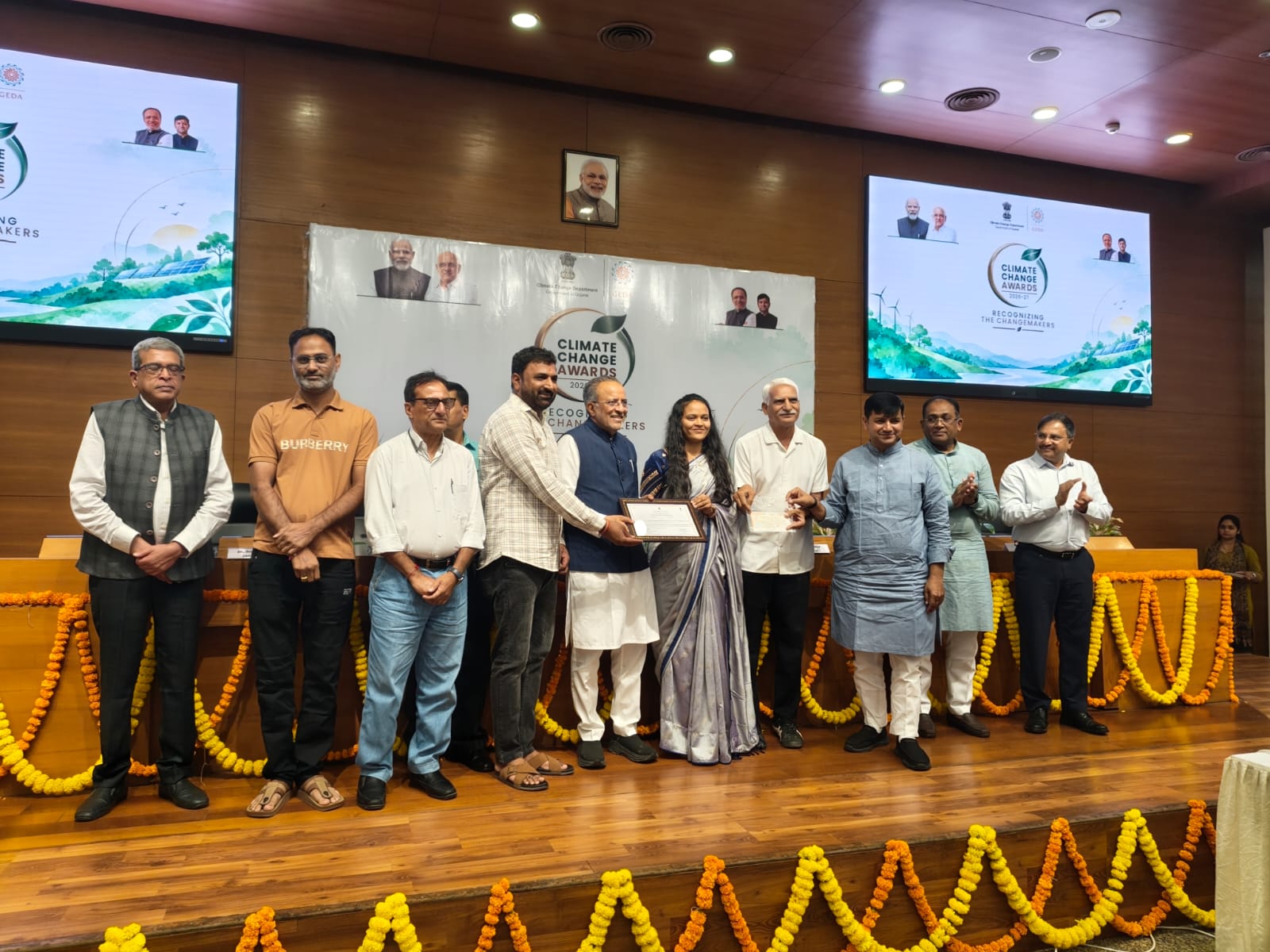 The award being presented on stage at the Climate Change Awards 2026-27 ceremony in Gandhinagar, under screens reading “Climate Change Awards — Recognizing the Changemakers”, with Gujarat government and GEDA officials