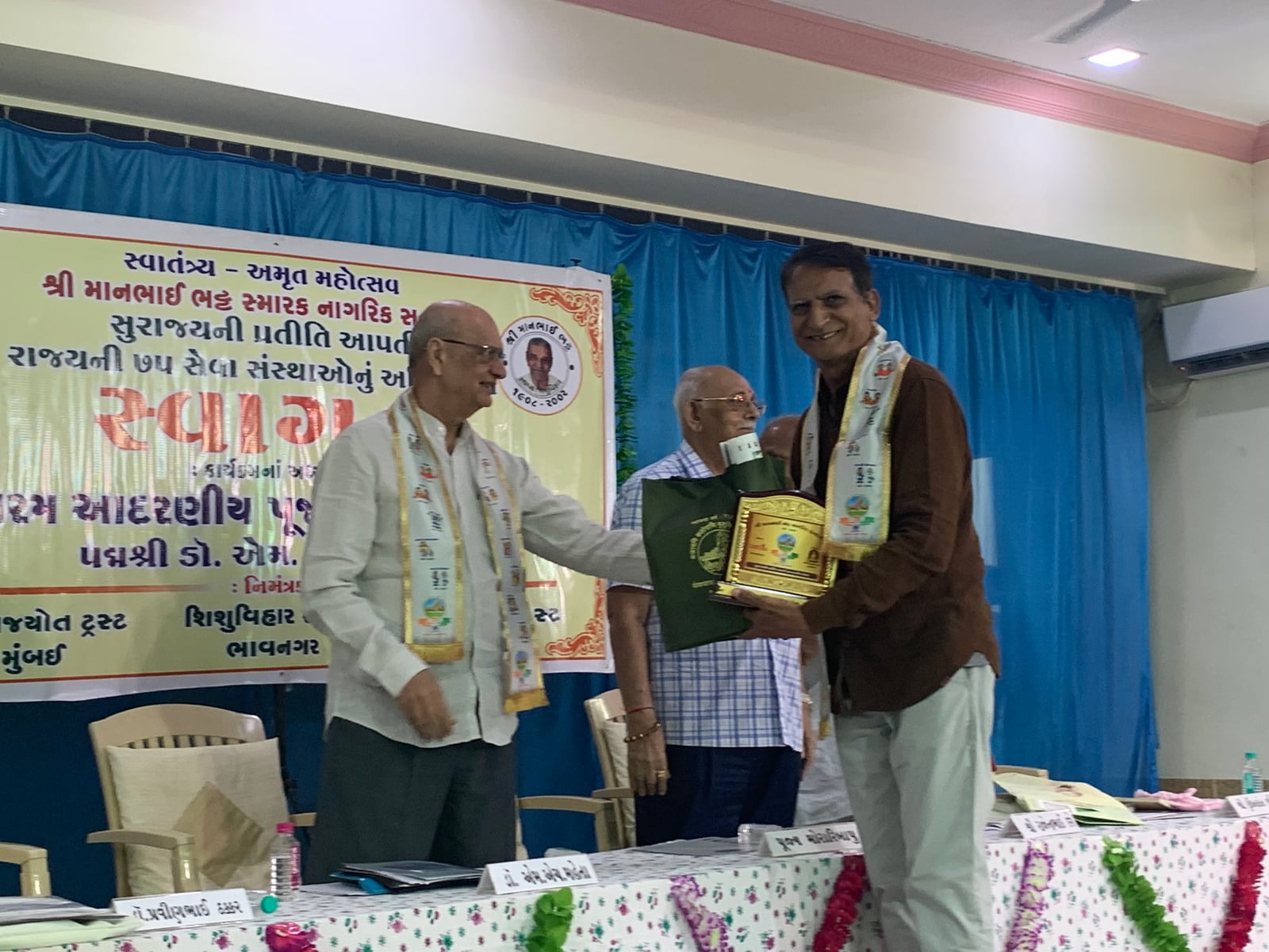 ભાવનગરમાં સમારંભના મંચ પર પુરસ્કાર અર્પણ થઈ રહ્યો છે; પાછળના બેનરમાં રાજ્યની ૭૫ સેવા સંસ્થાઓનું સ્વાગત