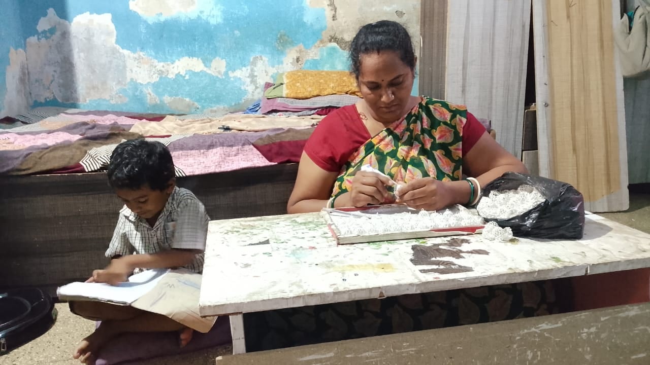 A woman threading imitation jewellery at a low table on the floor, her young son doing his schoolwork at the other end of the same table