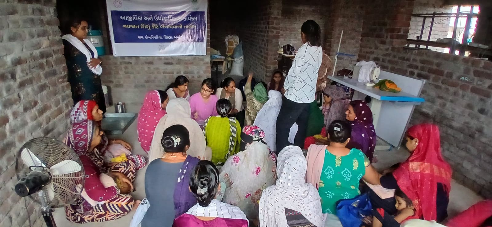 About twenty-five women seated on the floor of a brick training room watching a demonstration, sewing machines along the far wall, under a banner naming the training and the village