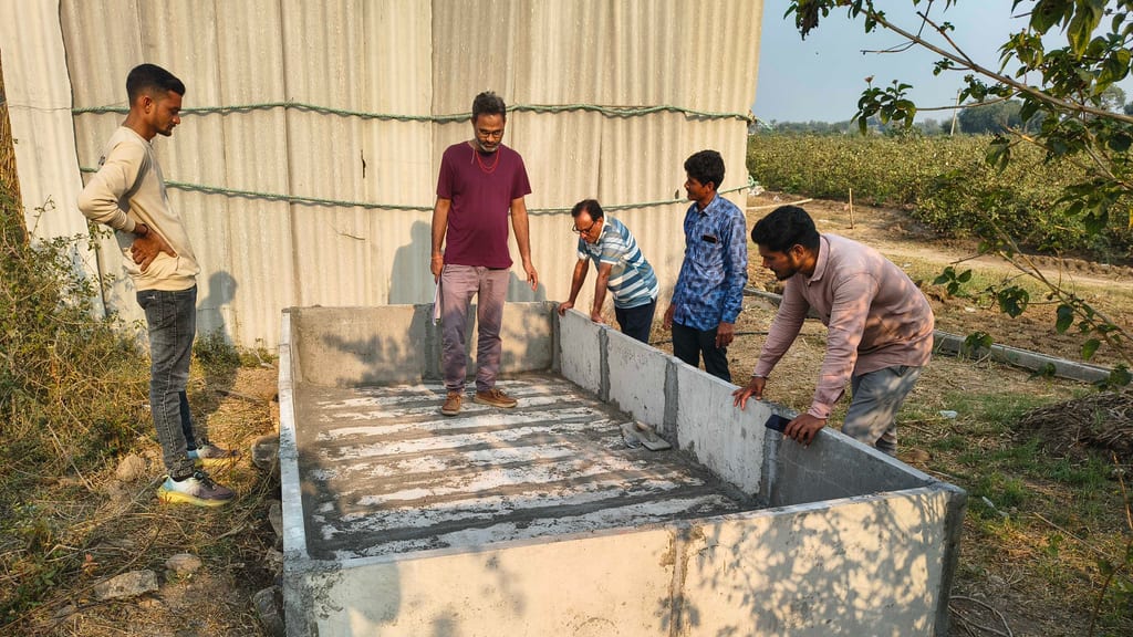 પતરાના શેડ પાસે નવો બંધાયેલો કોંક્રીટનો વર્મીકમ્પોસ્ટ ટાંકો, અને પૂરી થયેલી દીવાલો તપાસતા પાંચ પુરુષો