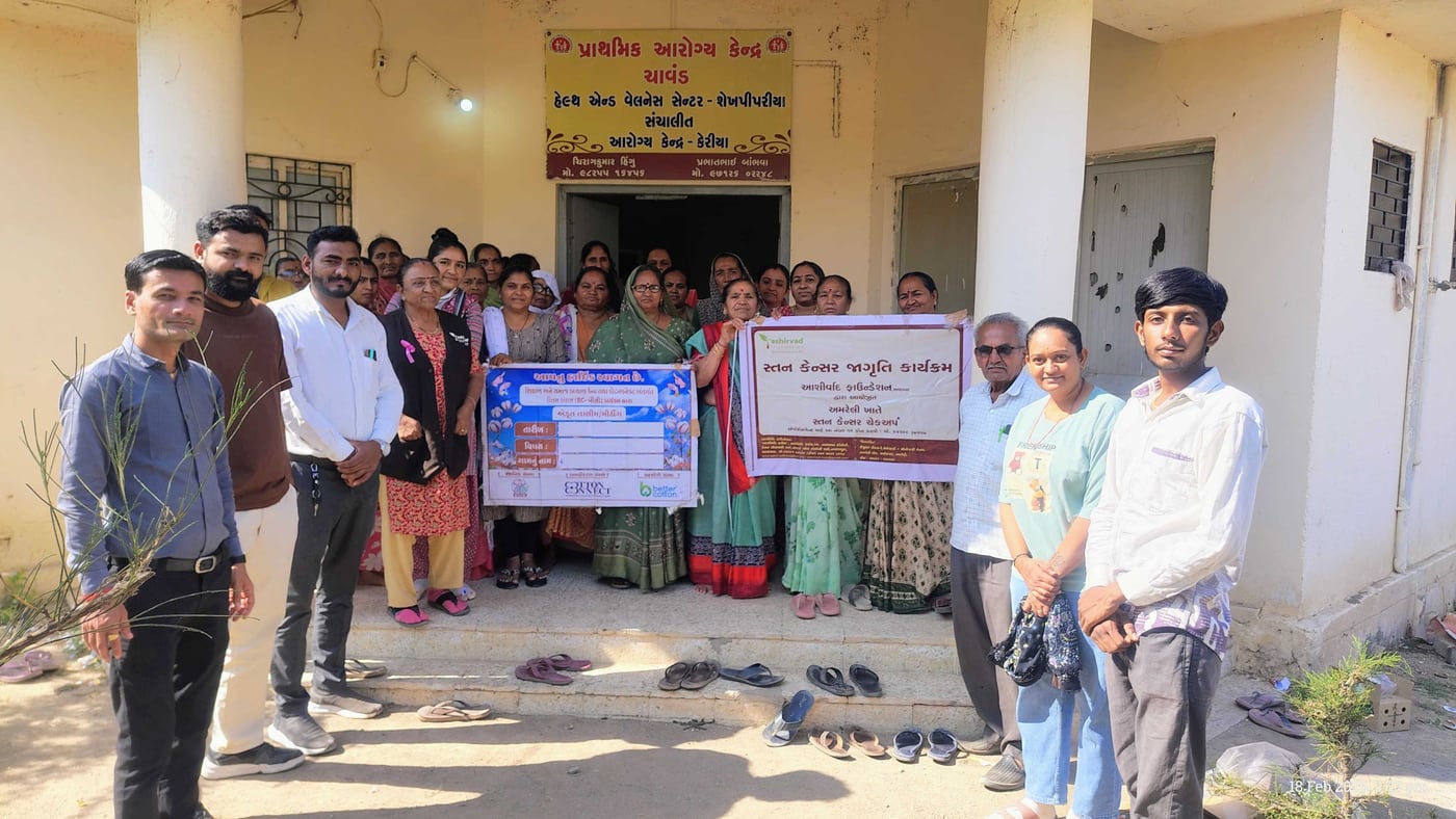 A group of women and staff outside a primary health centre, holding a banner reading “સ્તન કેન્સર જાગૃતિ કાર્યક્રમ” — breast cancer awareness programme