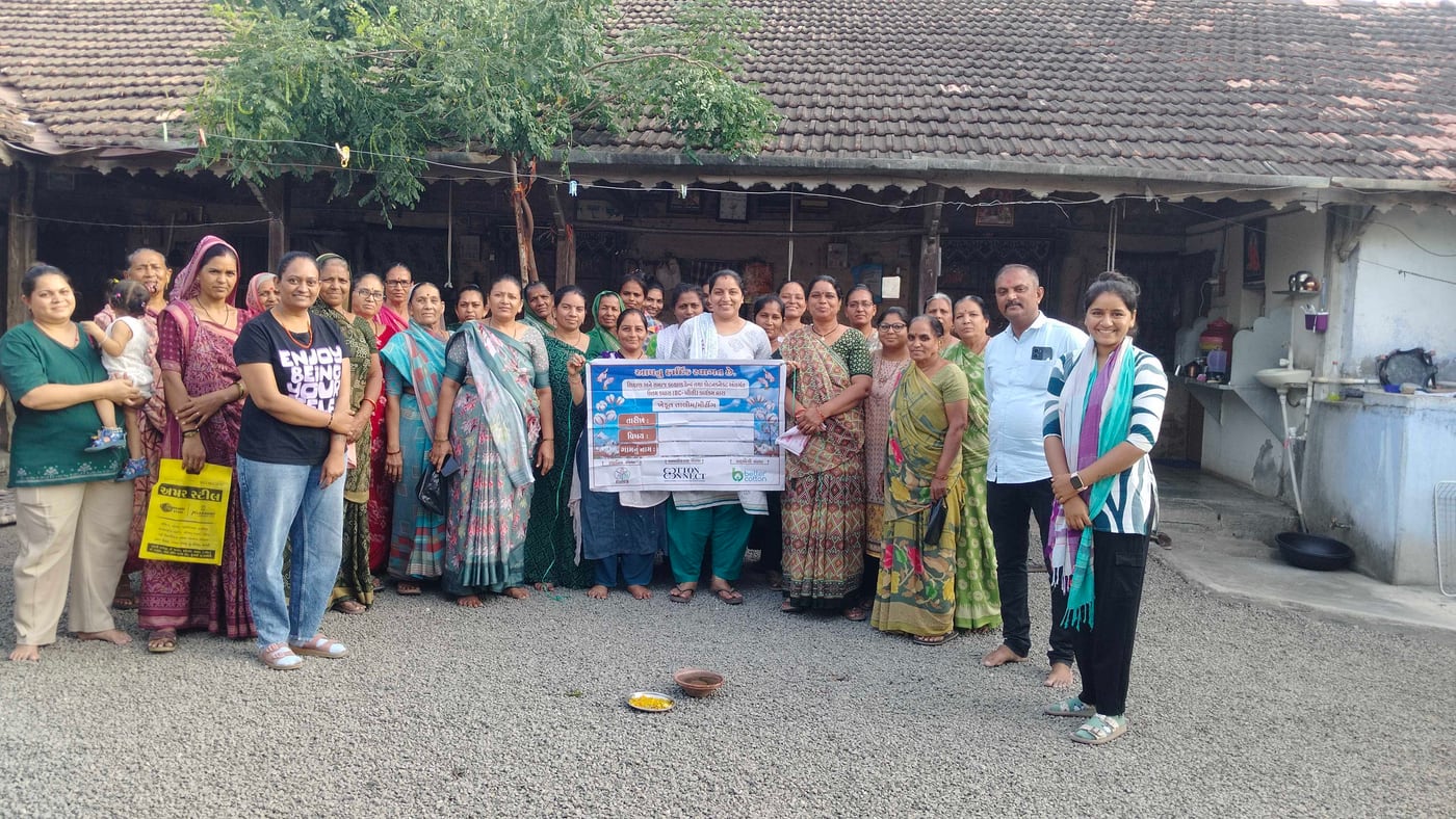 A large group of women standing in a village courtyard with two SSKK staff, holding a printed banner