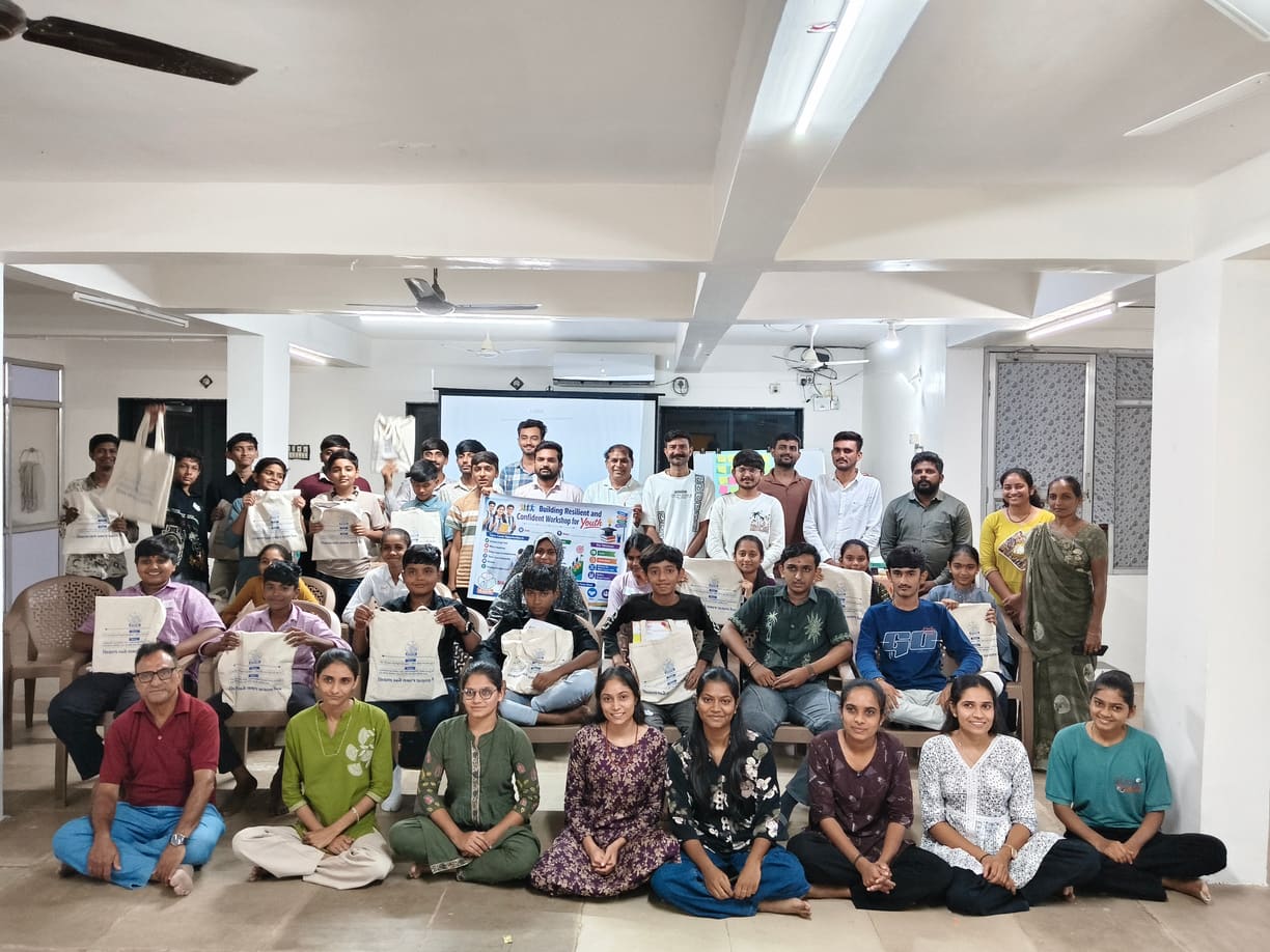 Youth Workshop participants and facilitators sitting together for a group photo, several holding tote bags, after the final session at the Head Office in Amreli